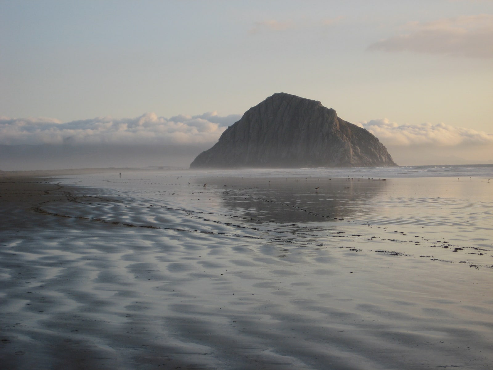 Morro Rock reflected in rippled wet sand and backed by fluffy white clouds on a dreamy evening.