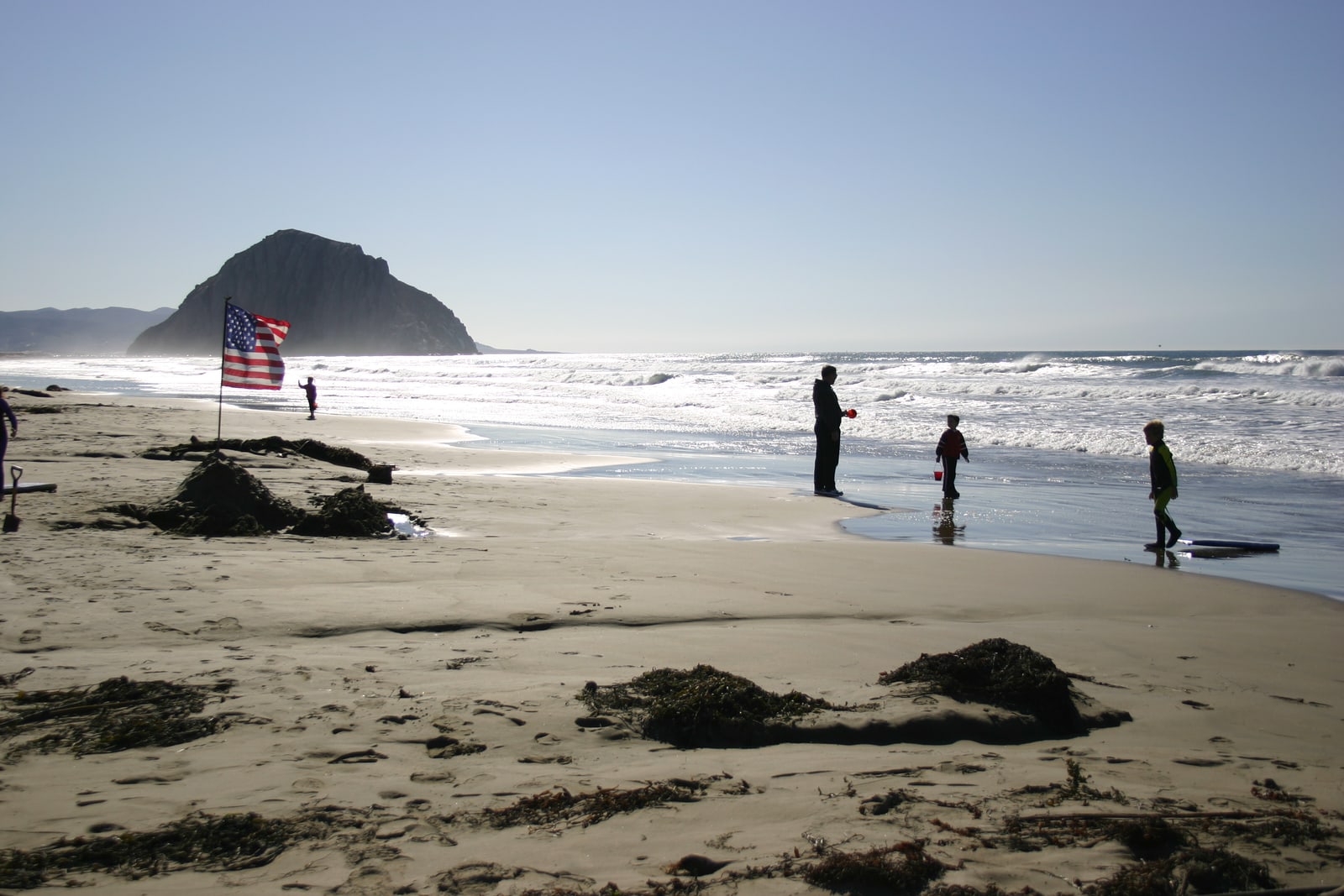 An American flag flies on the beach near Morro Rock while children and adults play in the water nearby.