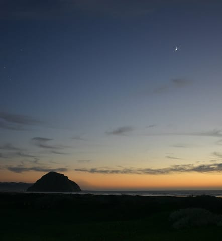 A crescent moon hangs in the night sky above Morro Rock. 